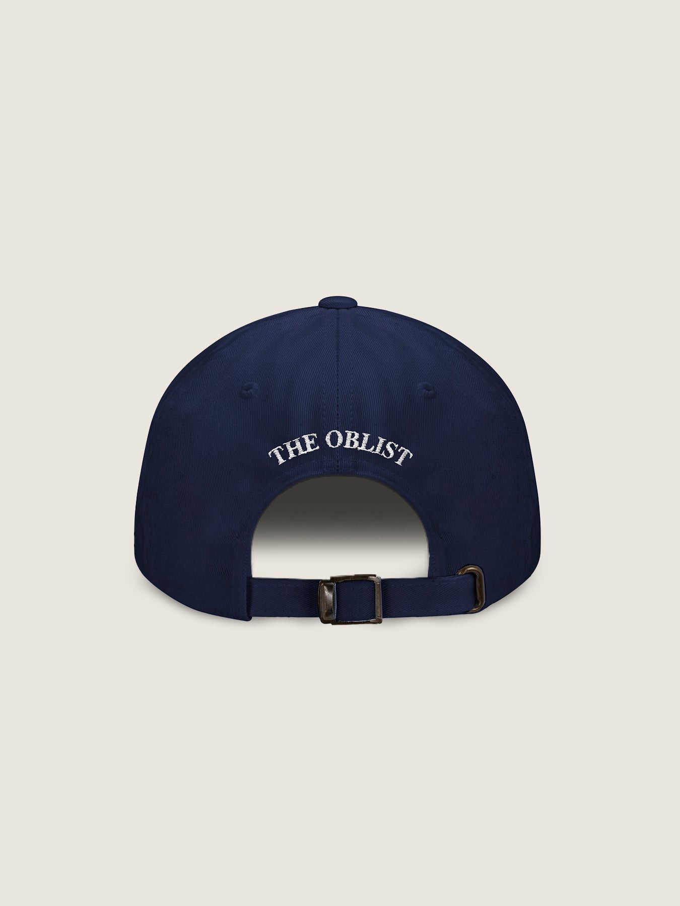 The image features the back of "The Curated Cap" by The Oblist, a navy blue baseball cap with an adjustable strap and metal buckle. "THE OBLIST" is elegantly embroidered in white above the opening. This limited-edition unisex accessory stands out against a plain beige background, making it a versatile addition to your wardrobe.