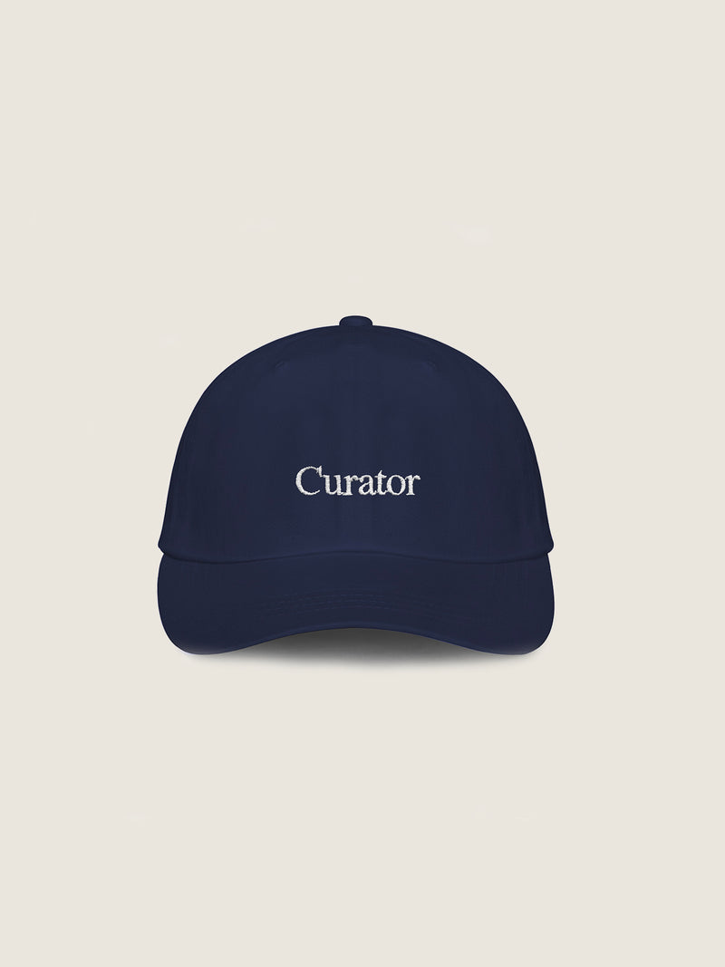 Introducing The Curated Cap by The Oblist, a navy blue baseball cap featuring "Curator" embroidered in white on the front. Crafted from breathable fabric for ultimate comfort, this unisex cap suits everyone and showcases a timeless design against its plain off-white background.
