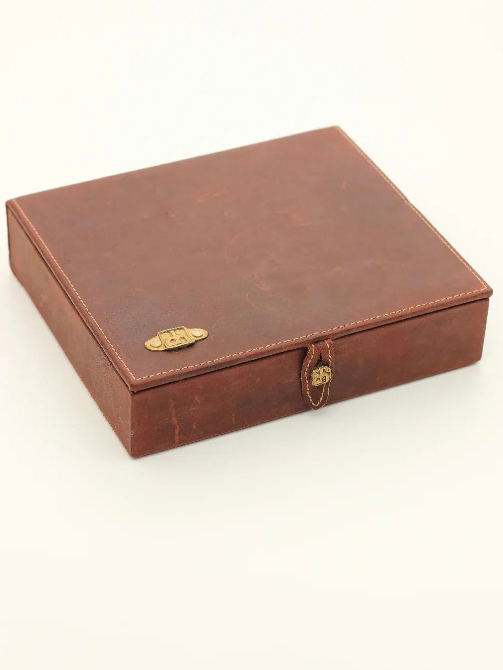 A vintage brown leather box, reminiscent of Boga Avante Shop's English Solitaire design, features a small gold-colored clasp that brings to mind David Marshall's creations. Its slightly worn texture suggests age or frequent use, hinting at the possibility of a cherished English Solitaire inside. The box sits elegantly on a plain, light-colored surface.