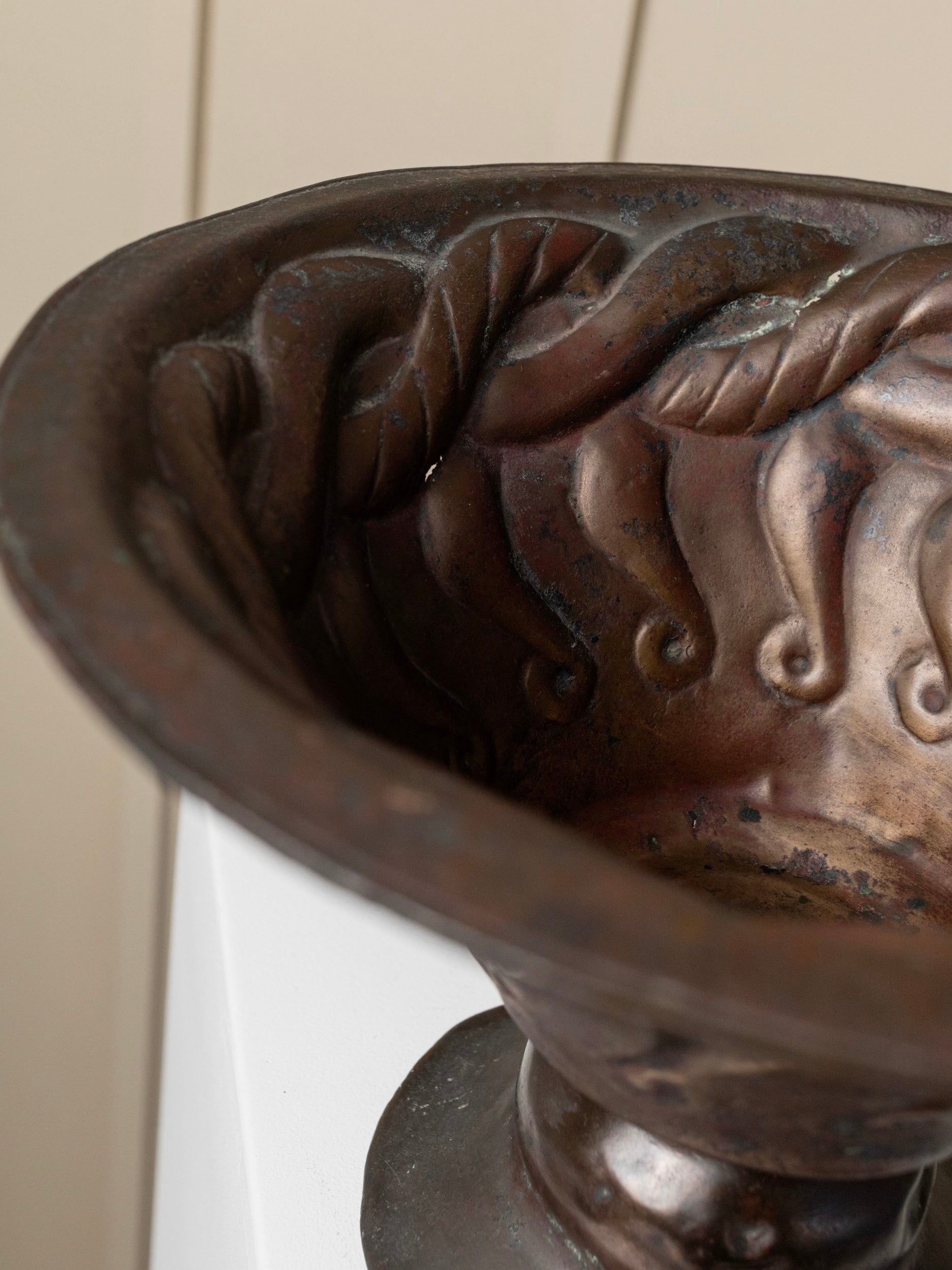 A close-up of the Bottega Jacobs Antique Fruit Bowl - Hand-Carved Copper, featuring decorative swirls and intricately worked snake motifs, elegantly resting on a pedestal. The patina finish enhances its aged and artistic design details, set against a softly blurred background.