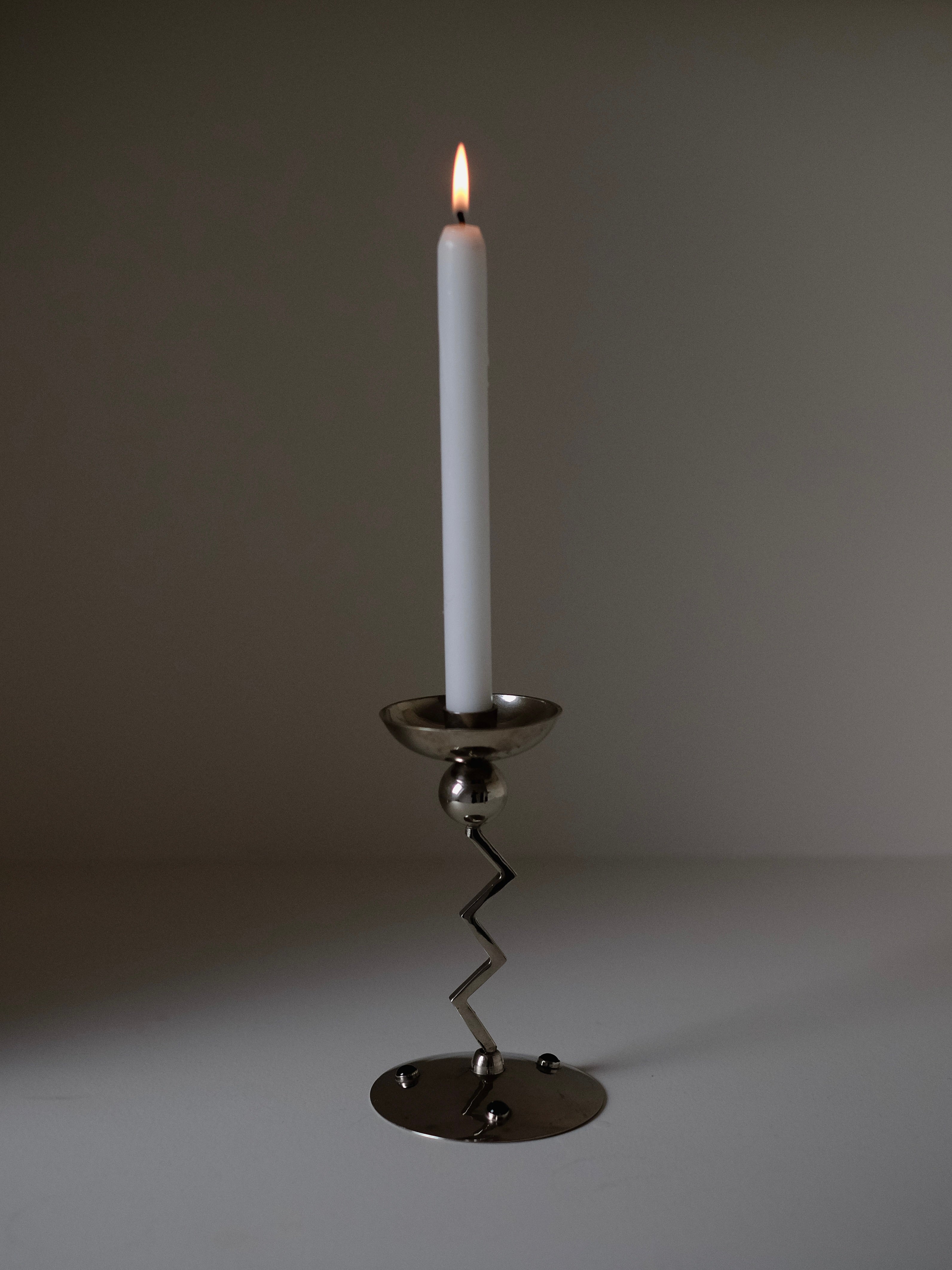 A lit white candle rests in a septembre studios Zigzag Candleholder with a zigzag stem, channeling an 80's aesthetic. Positioned on a flat surface against a plain backdrop, it casts soft shadows while its flame gently illuminates the surroundings.