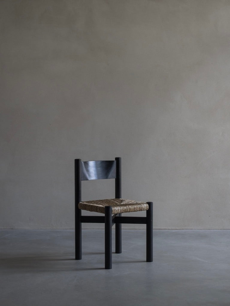 The 'Les Allues' Straw Chair by Bicci de' Medici, crafted from sleek mahogany with a woven seat, rests against a plain neutral wall on a bare concrete floor. This minimalist setting enhances the exquisite craftsmanship of the chair's design.