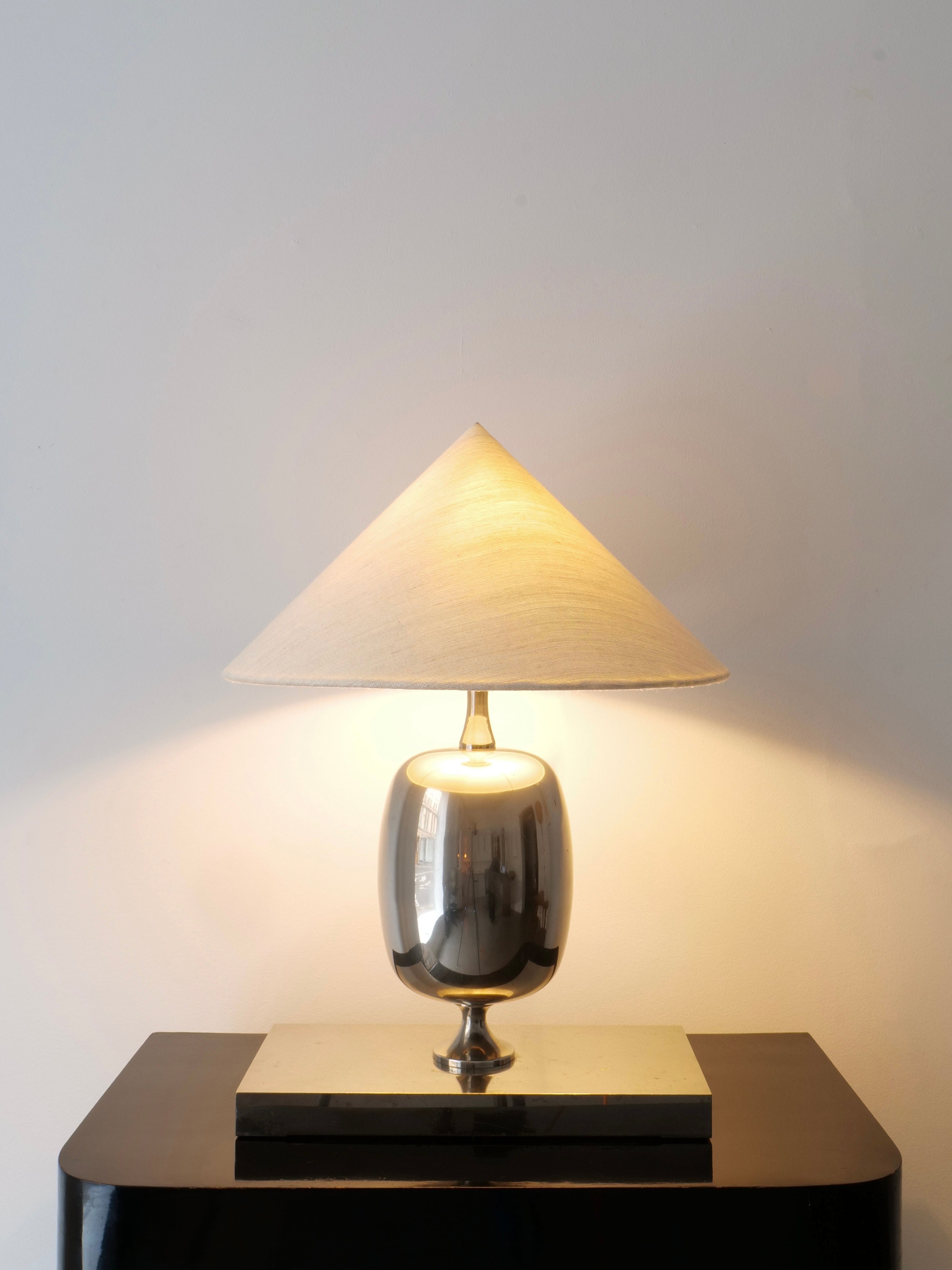 The Barbier Chrome Table Lamp by Collection Apart, featuring a conical beige shade and chrome over brass design, is placed on a sleek black surface. It emits a warm light that softly illuminates the neutral-colored wall behind it.