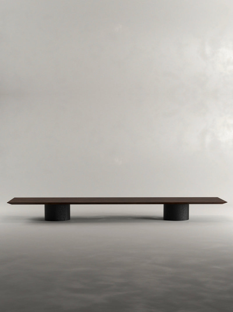 The 'Pier' Coffee Table by Siete Studio is a minimalist long rectangular piece, handcrafted from solid black oak with two cylindrical black supports, set against a plain light gray background.