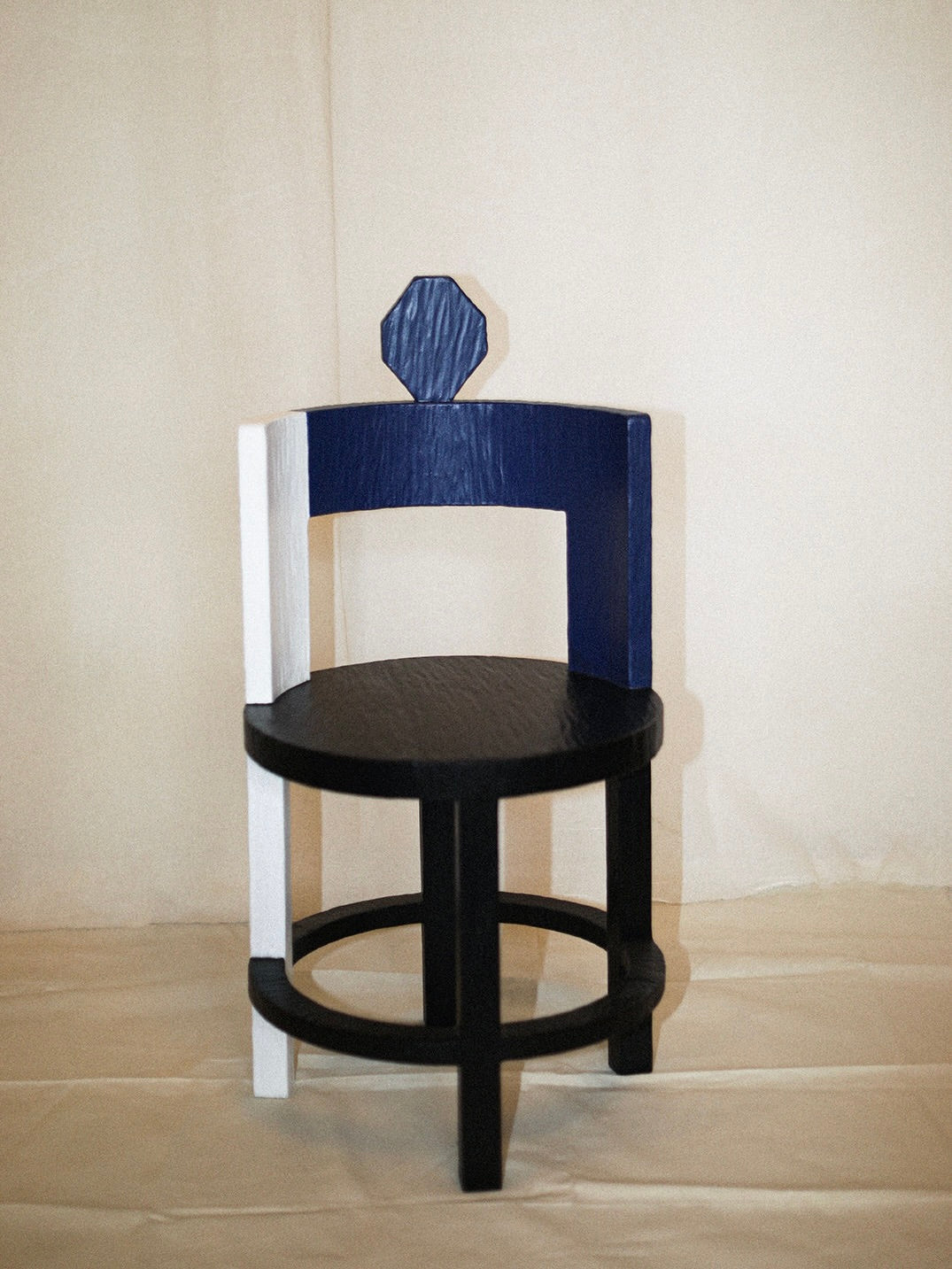 The Rooms Studio's Sculptural Chair I in Primary Colors features a round seat, three legs, and a black base with a dark blue backrest. It boasts a white vertical side accent and an octagonal top on the backrest, all crafted in reclaimed oak against a plain backdrop.