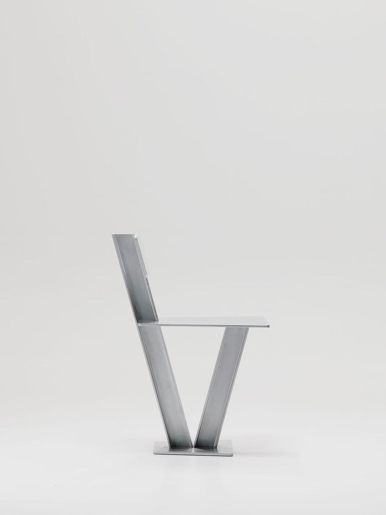 Side view of Marquel Williams' minimalist Beam Chair with a sleek V-shaped waxed aluminum frame, flat rectangular seat and backrest on a plain light gray background.