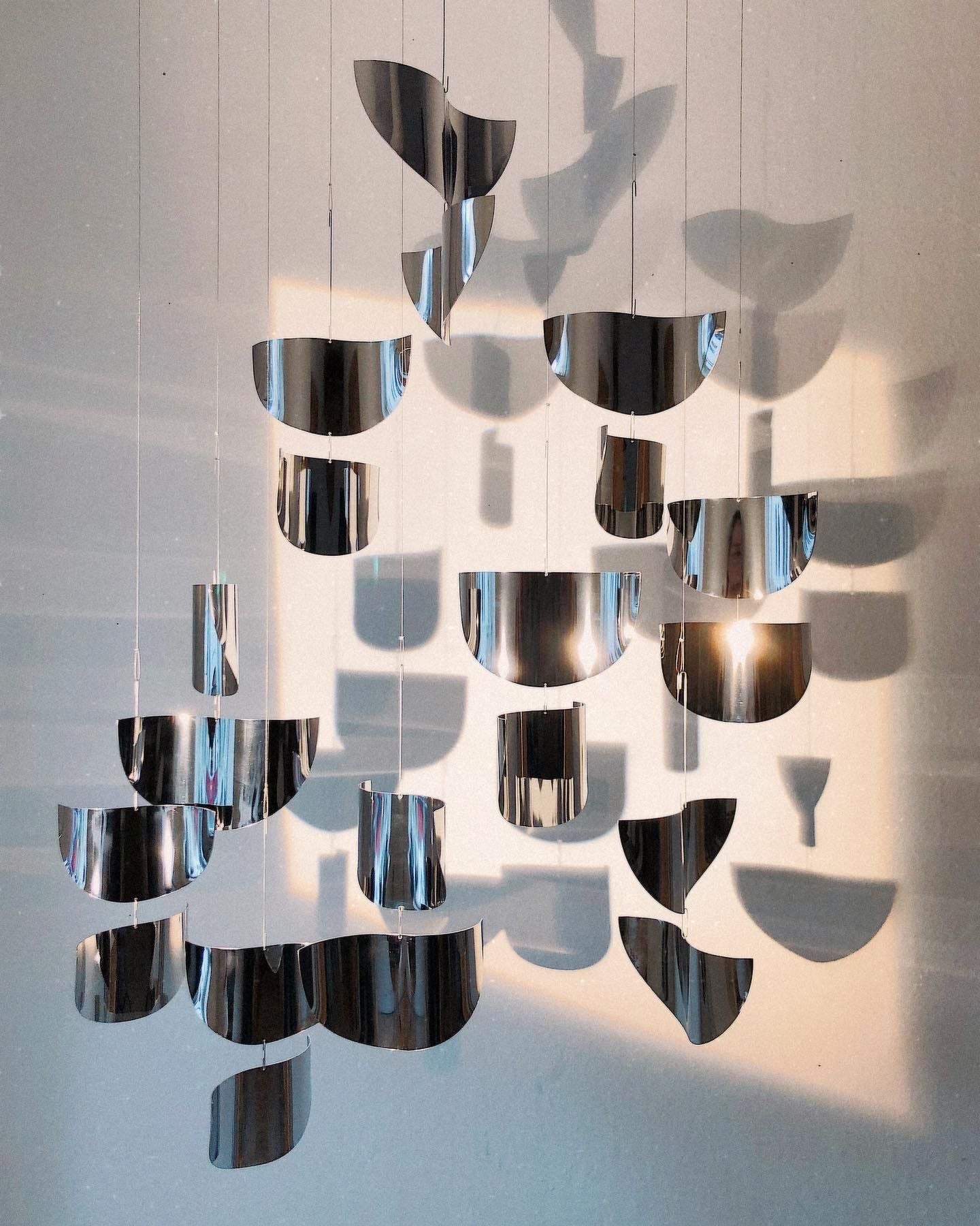 The Sun Catcher by Studio Fie Paarup is a modern, handmade Danish mobile with gleaming metallic shapes hung by fine wires, casting abstract shadows on light walls in soft natural light.