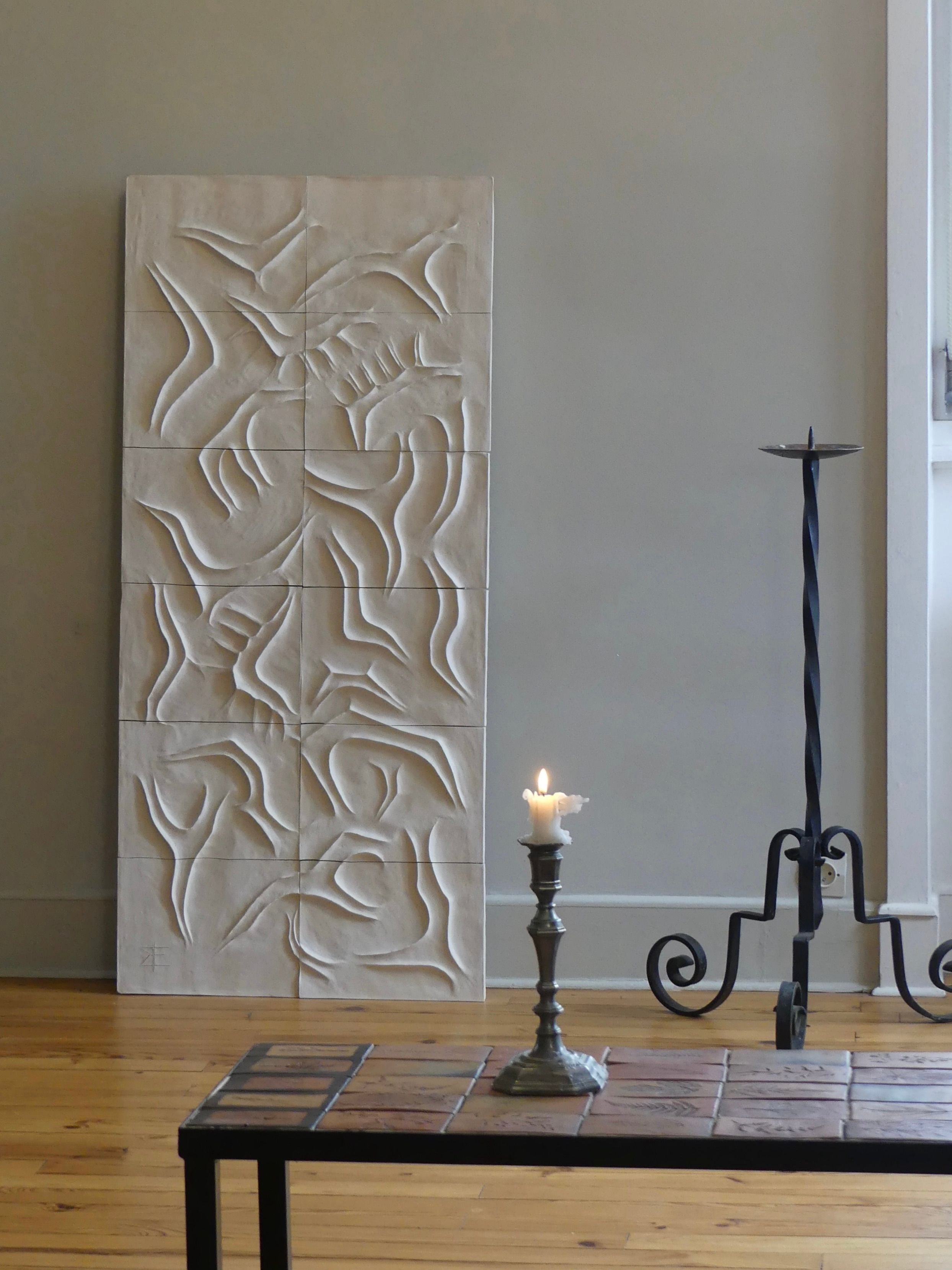 A Franck Scala "Scala Dei Turchi" decorative white bas-relief panel with abstract shapes leans against a light gray wall. In front, a lit white candle on a wrought iron holder stands beside a tall, twisted metal stand. The decor is finished by a wooden floor and tiled table echoing the sculpture design.