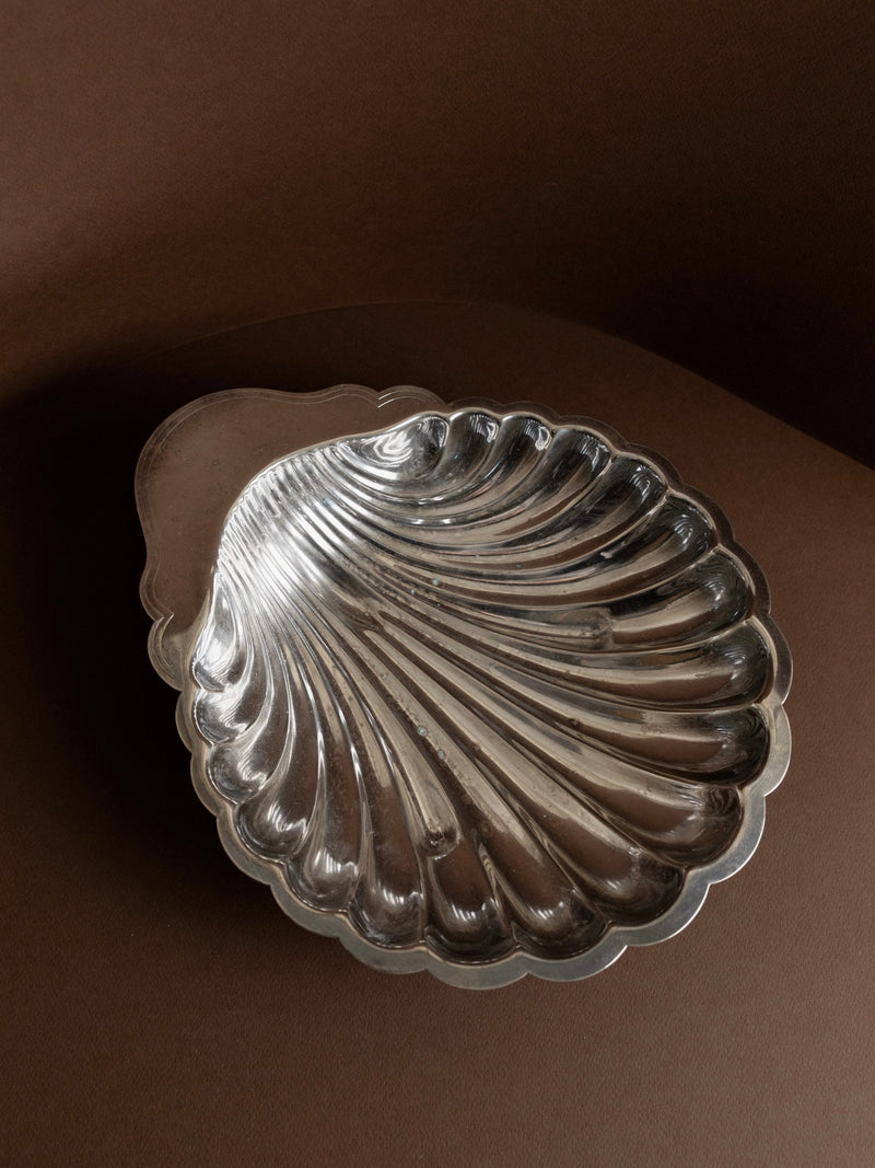 A Silver Plated Vintage Shell Shaped Bowl by Bottega Jacobs, expertly crafted for an Italian table decoration, gleams with a silver finish against a brown background. Positioned at an angle, its reflective surface accentuates intricate ridges, creating a stunning contrast with the matte backdrop.
