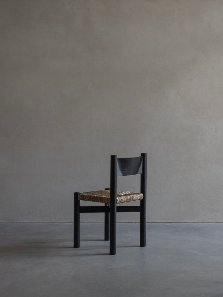 The 'Les Allues' Straw Chair by Bicci de' Medici stands against a plain gray wall on a smooth floor, its minimalist wooden frame and woven seat highlighted by soft lighting, showcasing its clean lines and expert craftsmanship.