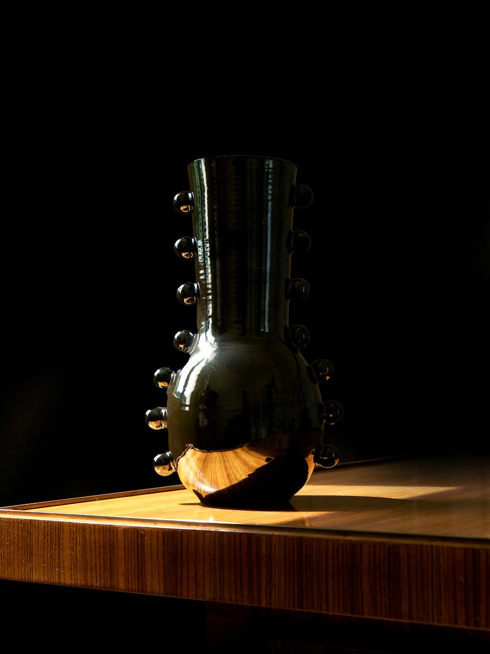 A wooden table is graced by the Vase Solstice II, a creation by Landy Rakoto Ceramics. This tall, glossy black ceramic piece features sculptural elements and round protrusions. It reflects light to create a metallic effect finish, highlighting its sleek form against a dark background.