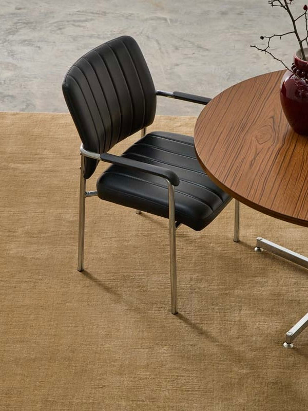 A black padded chair with metal arms sits by a round wooden table topped with a red vase of bare branches, all set on MOST’s 'Châtaigne' hand-woven rug in New Zealand wool.