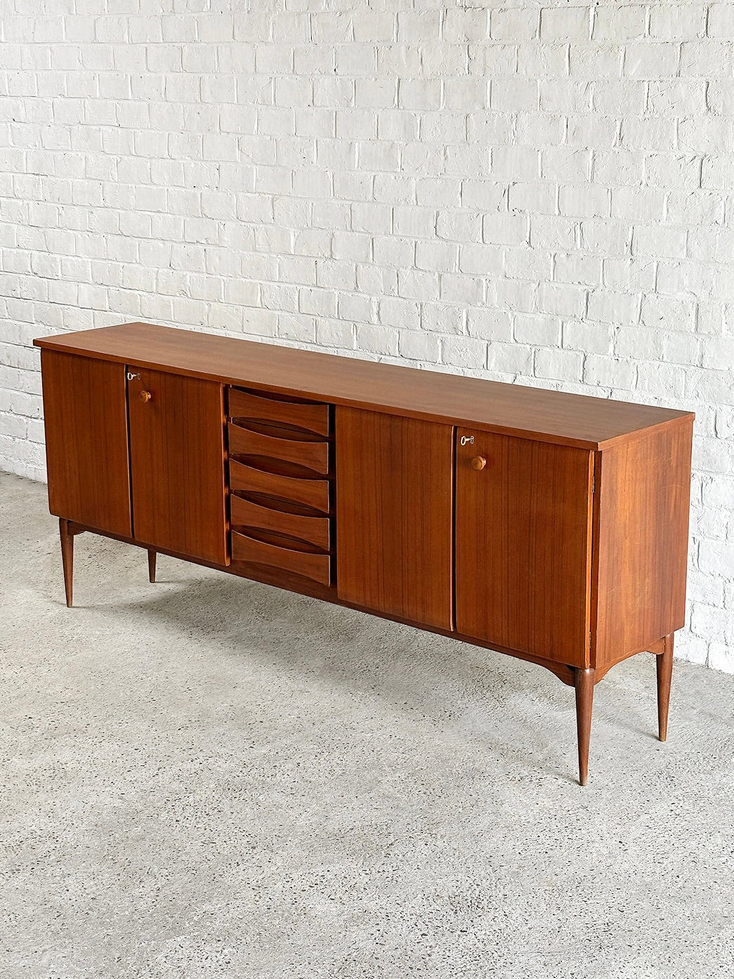 The Introverso Italian Modernist Teak Sideboard by Vittorio Dassi (1960's) features two cabinets and four central drawers with a warm wood finish. It stands on tapered legs, embodying mid-century charm against a white brick wall on a concrete floor.