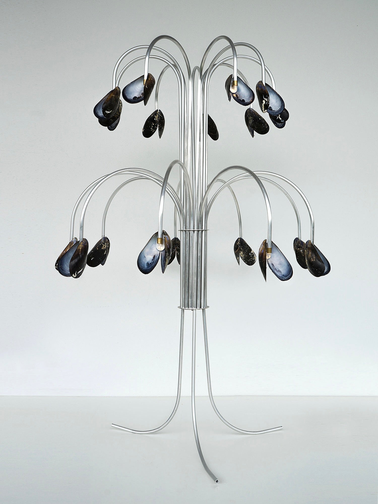 The Shell Light by Touch With Eyes is a whimsical sculpture resembling a metallic tree with curved branches, each ending in a blue-black mussel shell adorned with hints of mother-of-pearl. It stands on a minimalist, tripod-like base against a neutral background, exuding an aura of handcrafted elegance.