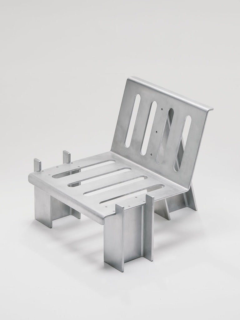 The Marquel Williams Beam Lounge Chair features a minimalist design with waxed aluminum, a low angular build, and a slatted backrest and seat on a strong geometric frame, all set against a plain, light background for a sleek modern look.