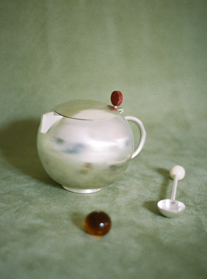 A shiny silver-plated Natalia Criado Teiera with a round body and a red knob on the lid, accompanied by a silver spoon, on a textured green surface. Nearby, two small round objects rest on the