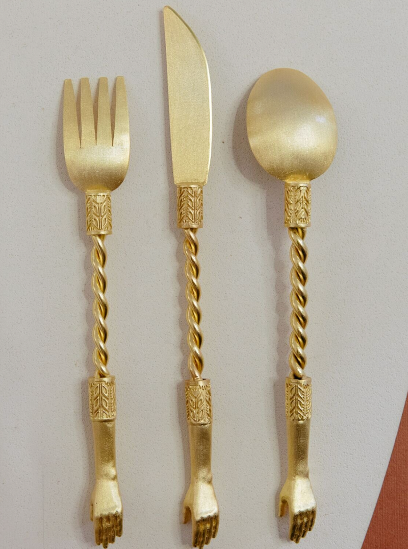 The Natalia Criado Hand Set features a trio of gold-colored utensils—fork, knife, and spoon—with contemporary twisted handles and ornamental patterns culminating in sculpted hands, all handcrafted to elegance on a pristine white surface.