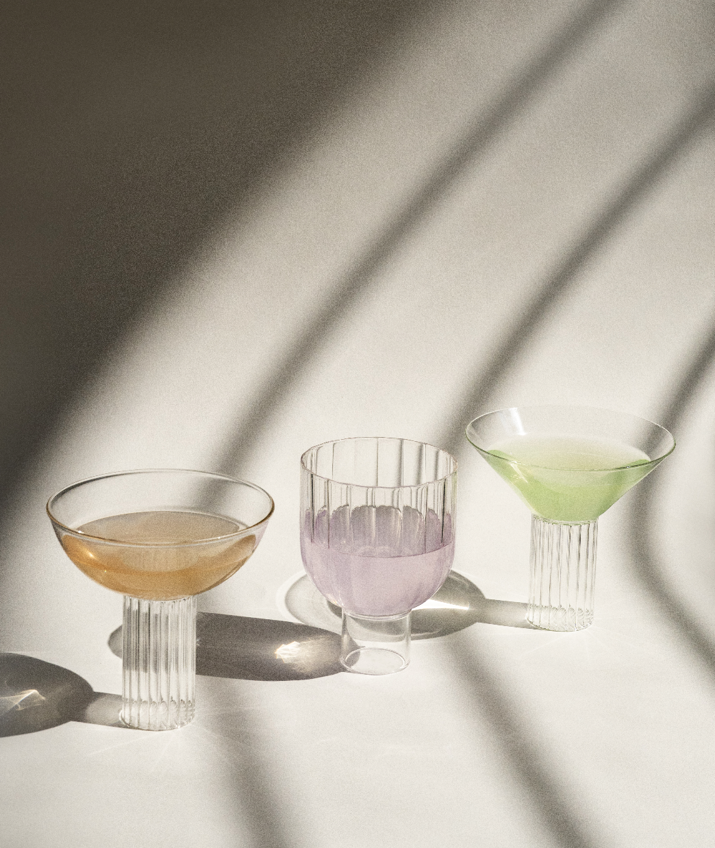 Three elegant Calici Milanesi cocktail glasses, forming a glassware trio with different colored drinks, illuminated by sunlight casting long shadows on a textured surface. (Agustina Bottoni)