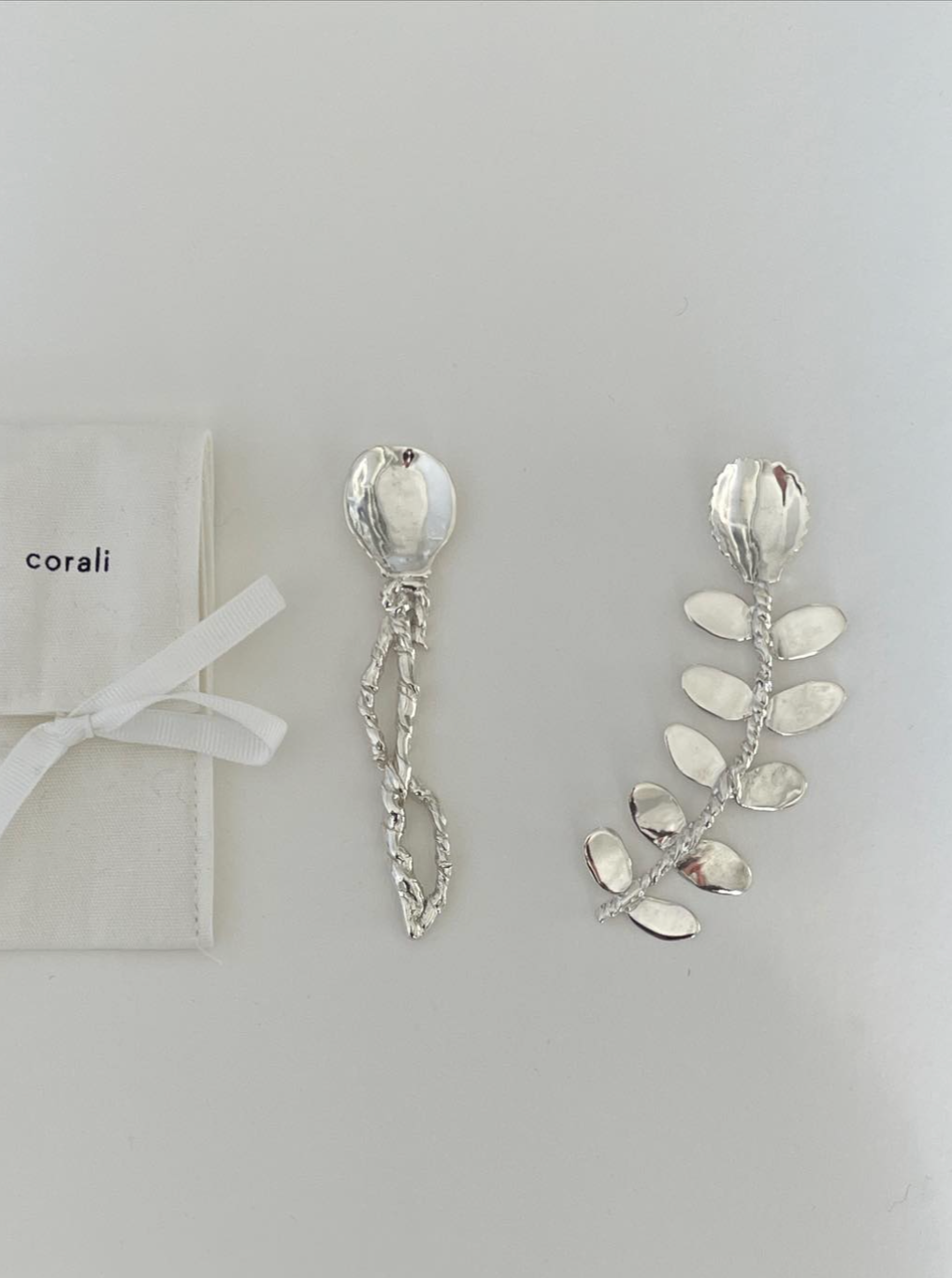 Two handcrafted silver hairpins with floral and leaf designs, displayed next to a small white pouch labeled "Babette Compote Spoon Sterling Silver" on a simple gray background from the Corali brand.