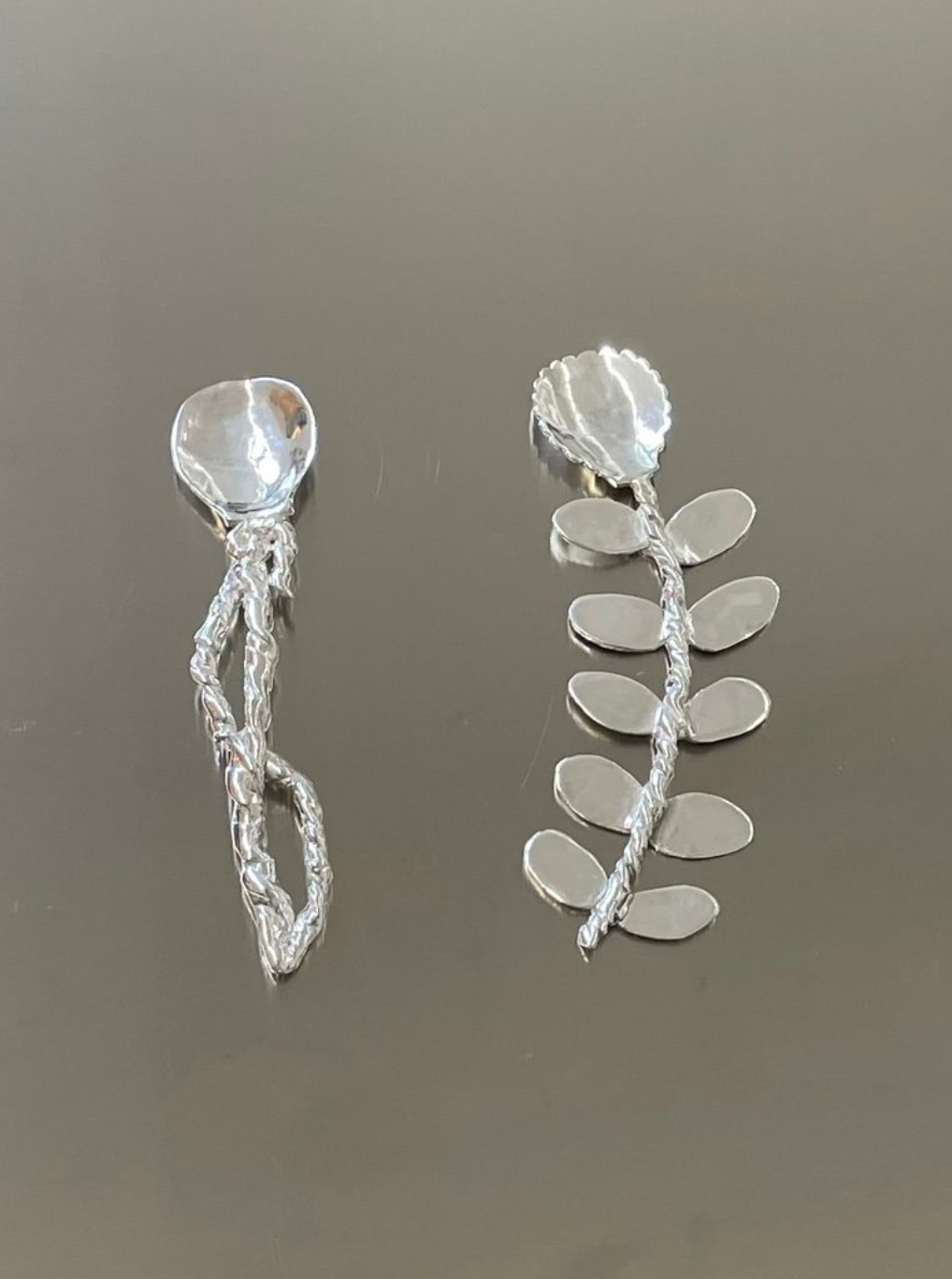 Two handcrafted Corali silver earrings with an organic design, featuring a textured twist and multiple leaf-like dangling elements, displayed against a grey background.