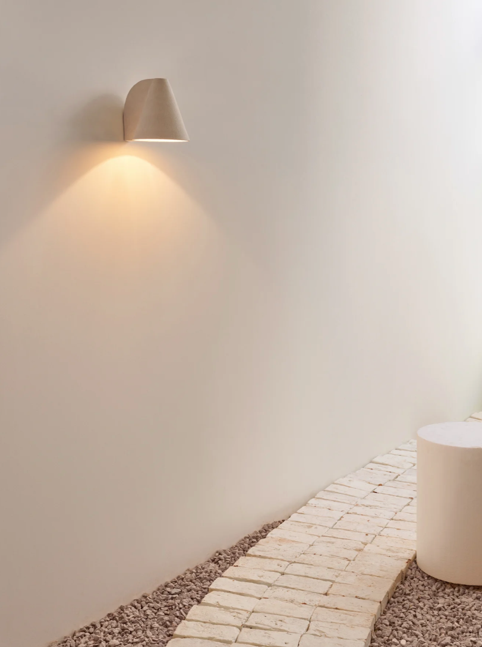 A corner wall features a Mariza Galani Esoteric Wall Lamp, emitting a soft, warm light above a textured stone pathway and a cylindrical white stool. The setting is minimalist and serene.