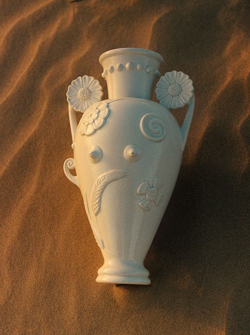 A White Earthenware Vase by Lobjet with intricate designs, including flowers and a snail shell, is placed on a sandy surface. Reminiscent of a Greek Amphora, the vase features two handles adorned with large, detailed flowers and a narrow neck that flares out at the top. The setting sun casts a warm glow.