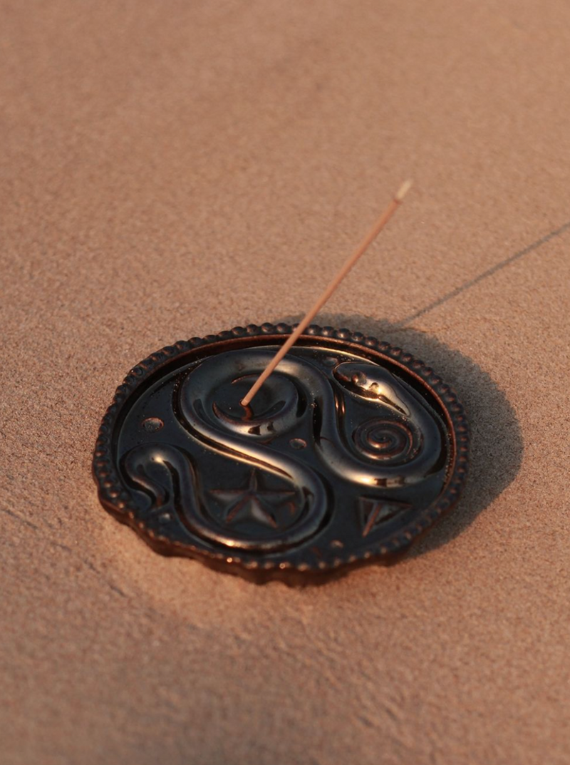 An Earthenware Incense Holder by Lobjet with a yin-yang design on a sandy surface conjures an air of serene home fragrance. A stick of incense stands upright in the center, reminiscent of ancient Greek amphora aesthetics. The sunlight creates a warm ambiance.