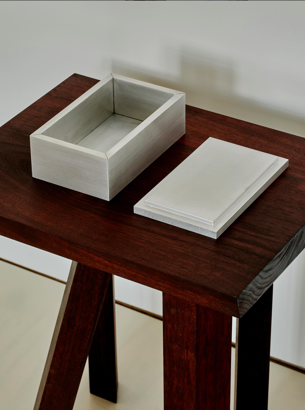 A small rectangular walnut Miter Box by MOCK STUDIO with a matching lid sits open on top of a dark wooden stool. The box's light color contrasts with the darker stool, which showcases visible wood grain and stands on a light-colored floor.