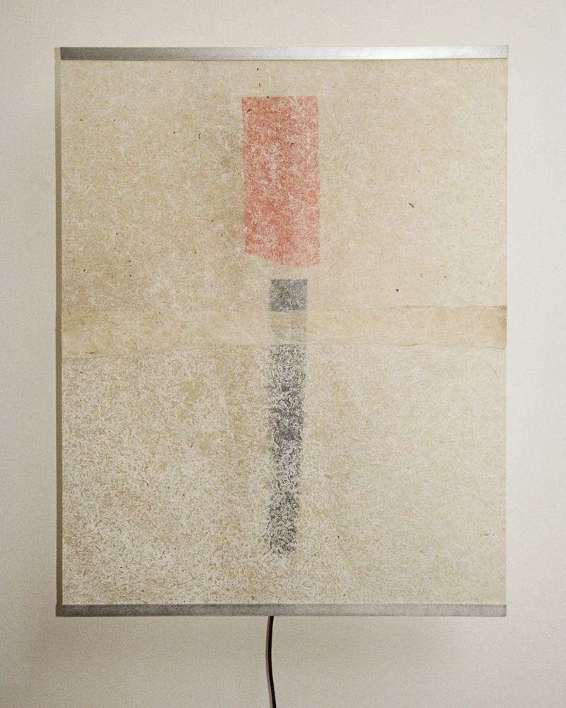 The Panel Lamp – Esei by TEGET features a textured design with a vertical red rectangle above a narrower black shape on beige, set in a metal frame and finished with a black cord, evoking the look of handmade art.