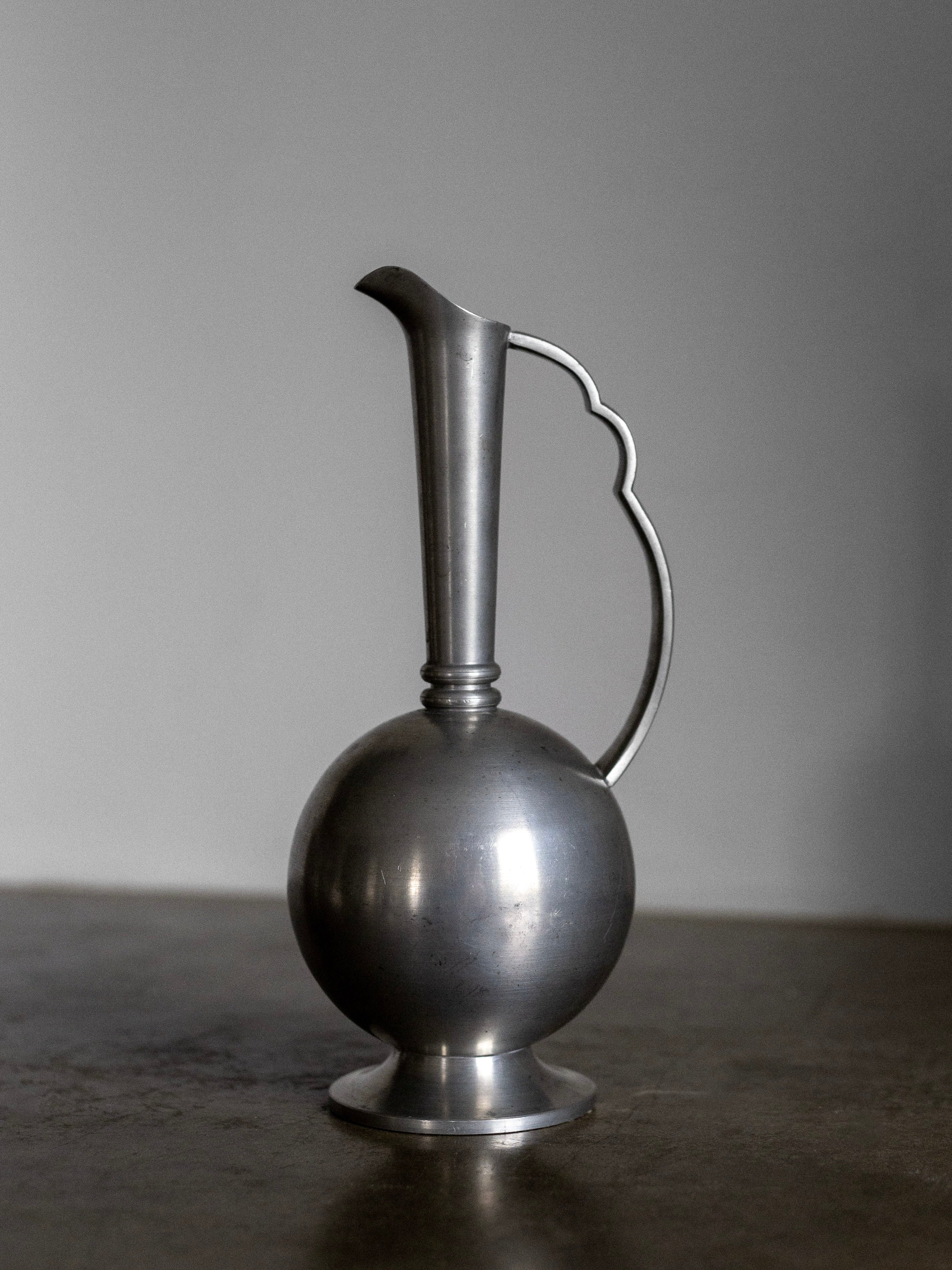 The Spigel Art Deco Metal Vase 1930s is a sleek, vintage-style piece with a spherical base and a curved, elongated spout. The handle boasts an artistic design with a delicate curve, exuding Swedish Art Deco charm reminiscent of CG Hallberg's creations. This collectible pewter vessel is beautifully showcased against a neutral gray background on a dark surface.