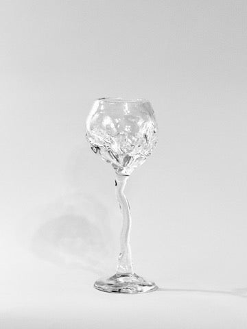A clear Hand Blown Freeform Wine Glass from Szkło Studio, crafted from transparent sodium glass, features a twisted stem and textured bowl. On a plain white background, lighting casts a faint shadow to the left, accentuating its unique design.