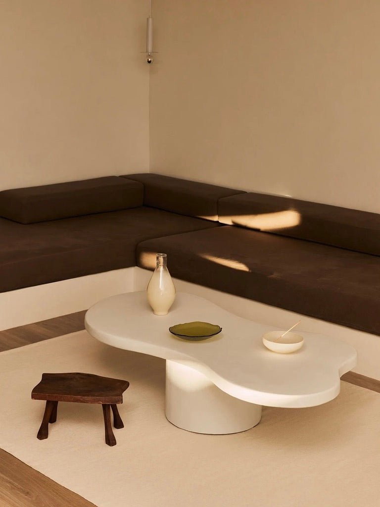 A minimalist living room is styled with a brown L-shaped sofa next to the MarlotBaus "Pesto" coffee table, crafted by artisans in a pristine white finish, accompanied by a small wooden stool. The coffee table is adorned with a bottle, a green plate, and a bowl with a spoon. Soft lighting enriches the cozy ambiance.