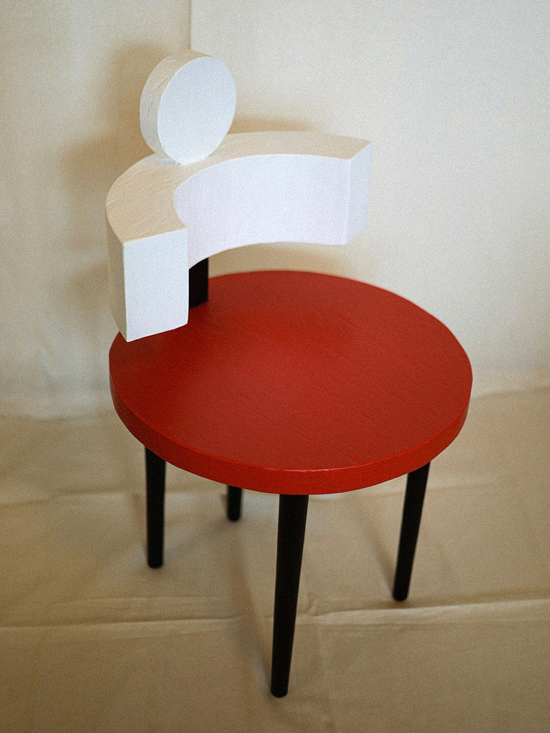 Rooms Studio's Sculptural Chair II in Primary Colors features a red circular seat, three black legs, and a white question mark-shaped backrest. Its design melds primary hues with artistic flair against neutral walls and flooring.