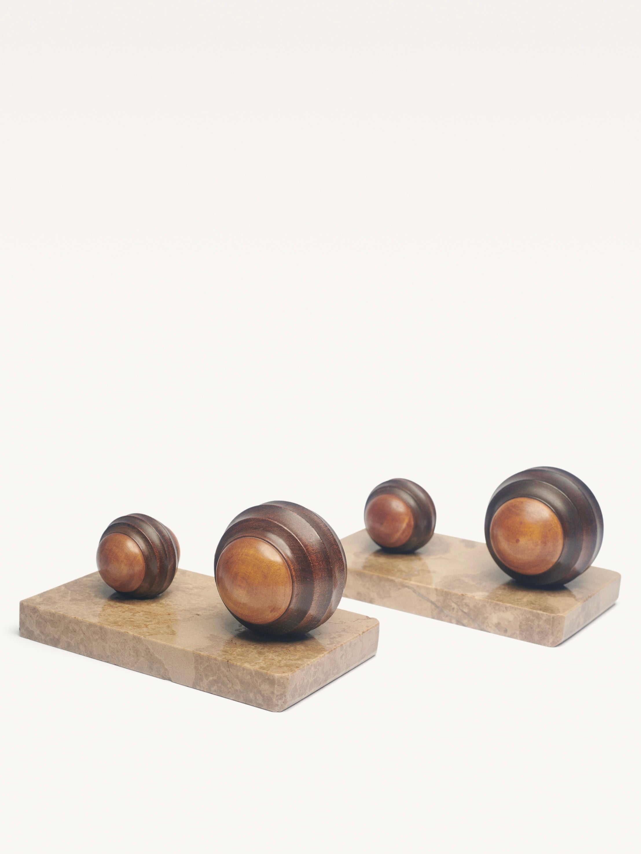 Four wooden spheres of varying sizes sit on two rectangular stone bases, blending modernist elegance and minimalism with their smooth, polished texture and rich brown tones. This pair of Art Deco Paper Press by Galerie MLS enhances the arrangement against a plain background.