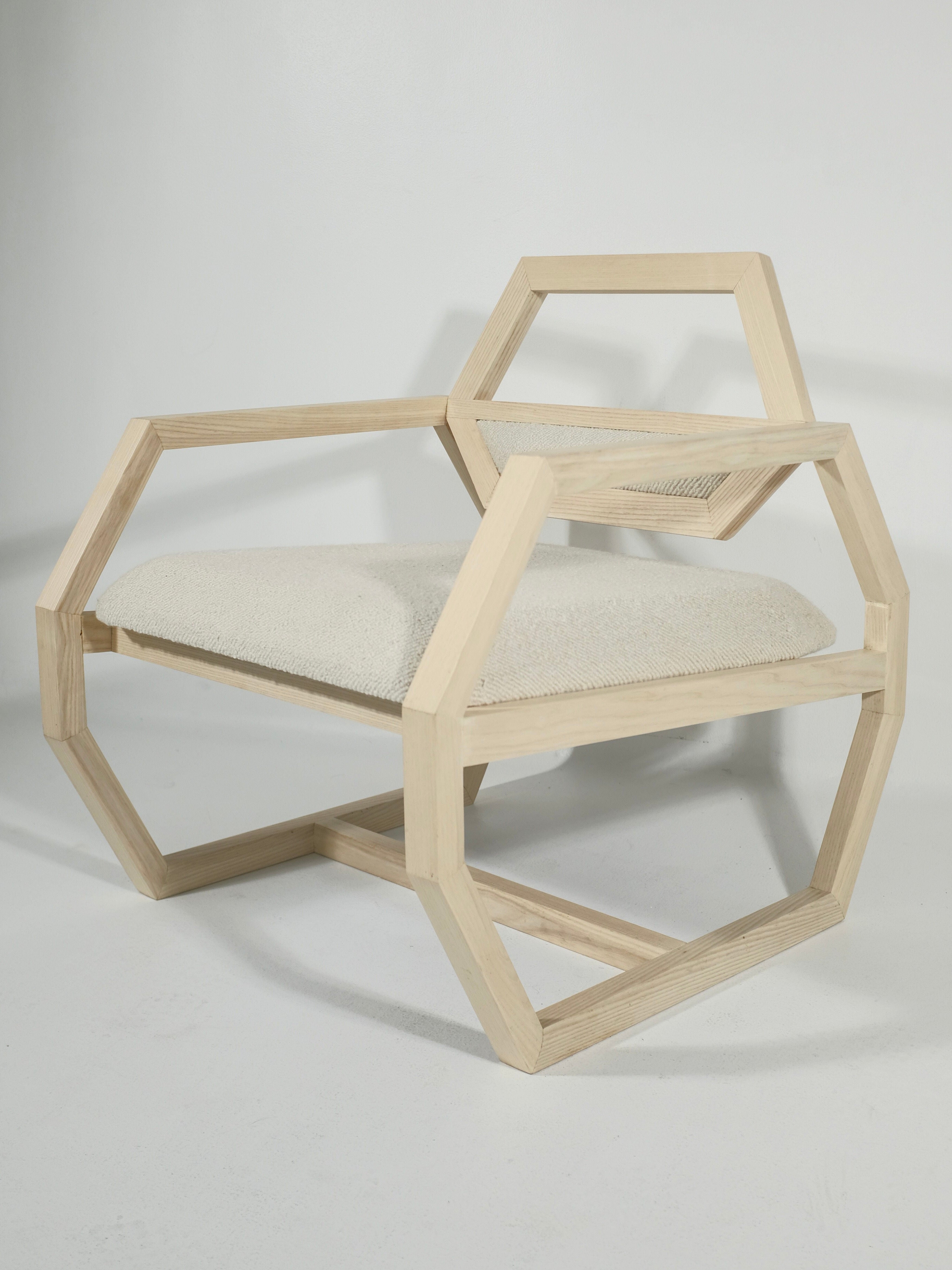 The Marcel Accent Chair by Nathan Chintala Design LLC is a modern, futuristic chair with a geometric design. It boasts a hexagonal frame complemented by a light beige cushioned seat. The open, angular structure features armrests and a back that continue the hexagonal motif, all set against a plain white background.