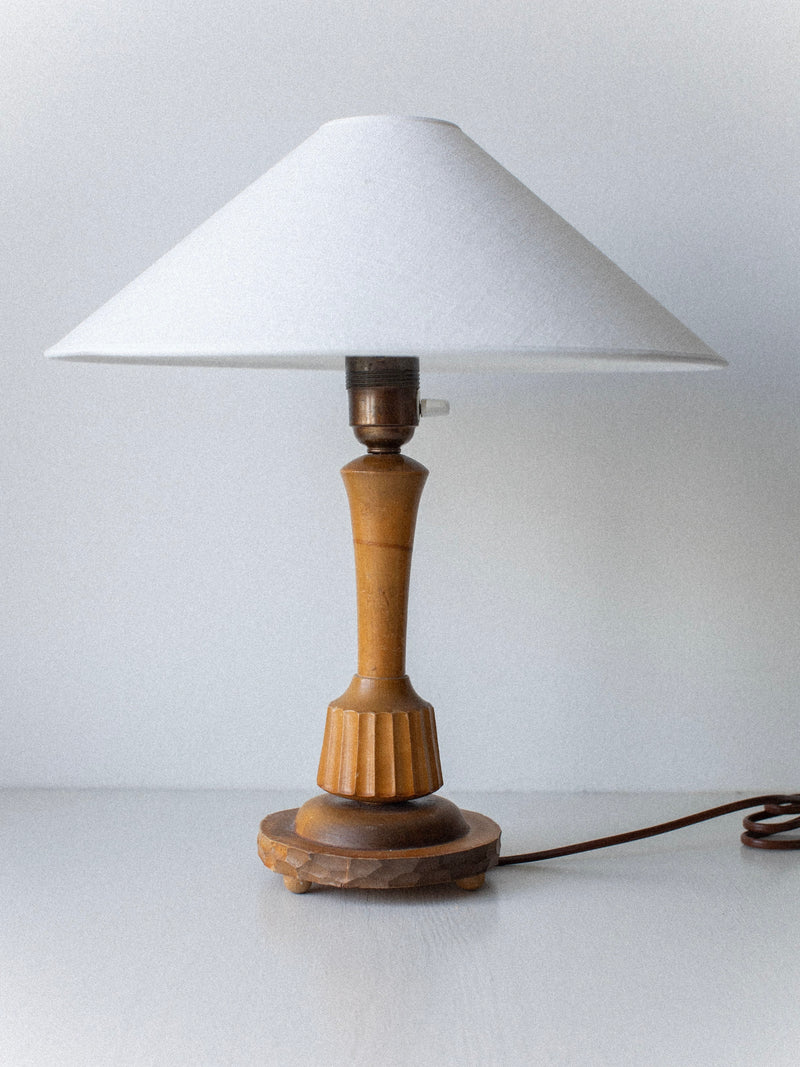The "1920's Table Lamp" by Out For Lunch, featuring a hand-carved wooden base and a white conical lampshade, stands against a plain, light-colored wall with its cord loosely coiled on the right side.