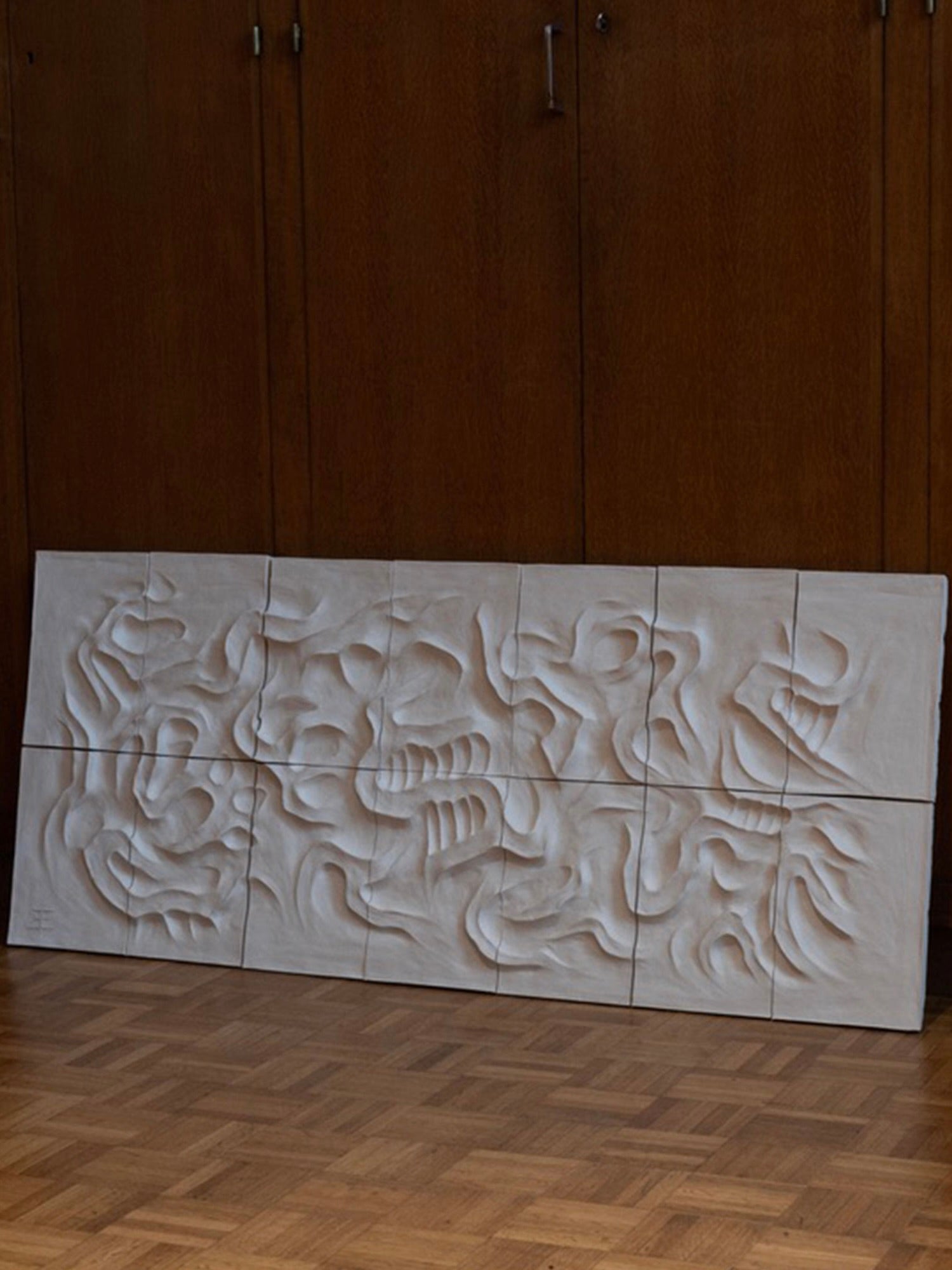 Le Grand Blanc - Wall Sculpture by Franck Scala features organic, flowing shapes in a light-colored, large-format ceramic bas-relief. Part of the RELIEFS collection, it stands against a wooden wall with wood-patterned parquet flooring below.