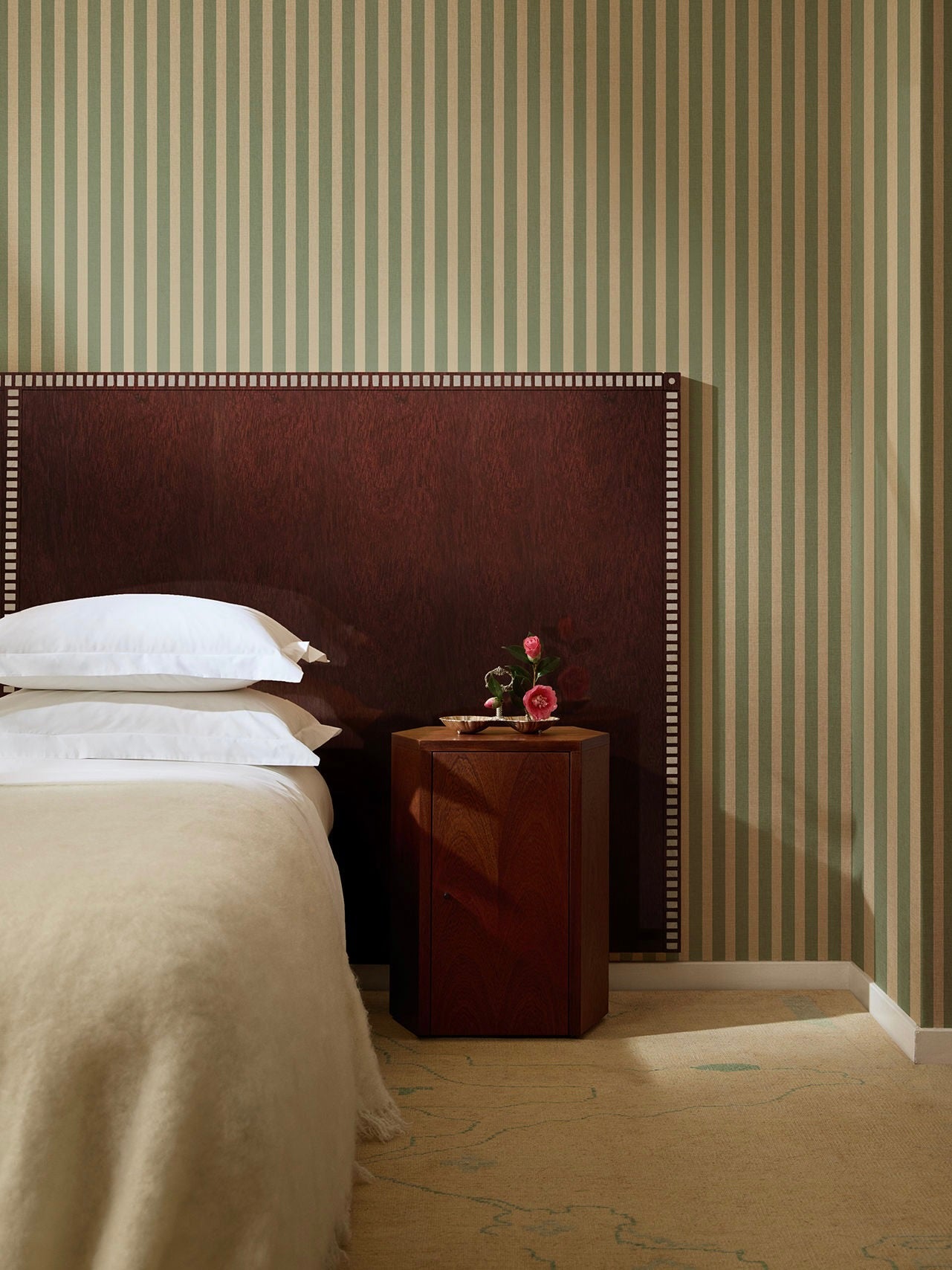 A cozy bedroom features a neatly made bed with white pillows and a beige blanket. The "Aquarama" Headboard in African Mahogany by Lemon is paired with vertical green and beige striped wallpaper. A wooden nightstand next to the bed holds a small floral arrangement in a vase.