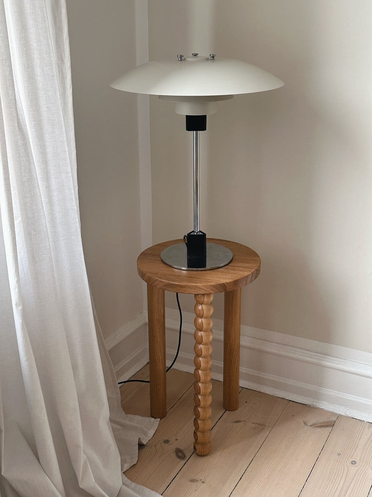 A modern white table lamp with a mushroom-shaped shade rests on the Piloti Side Table in Oak by Maitoinen Home, featuring twisted legs and an elegant Euclidean design. Situated in a corner next to white curtains, it's against a light-colored wall and hardwood floor.