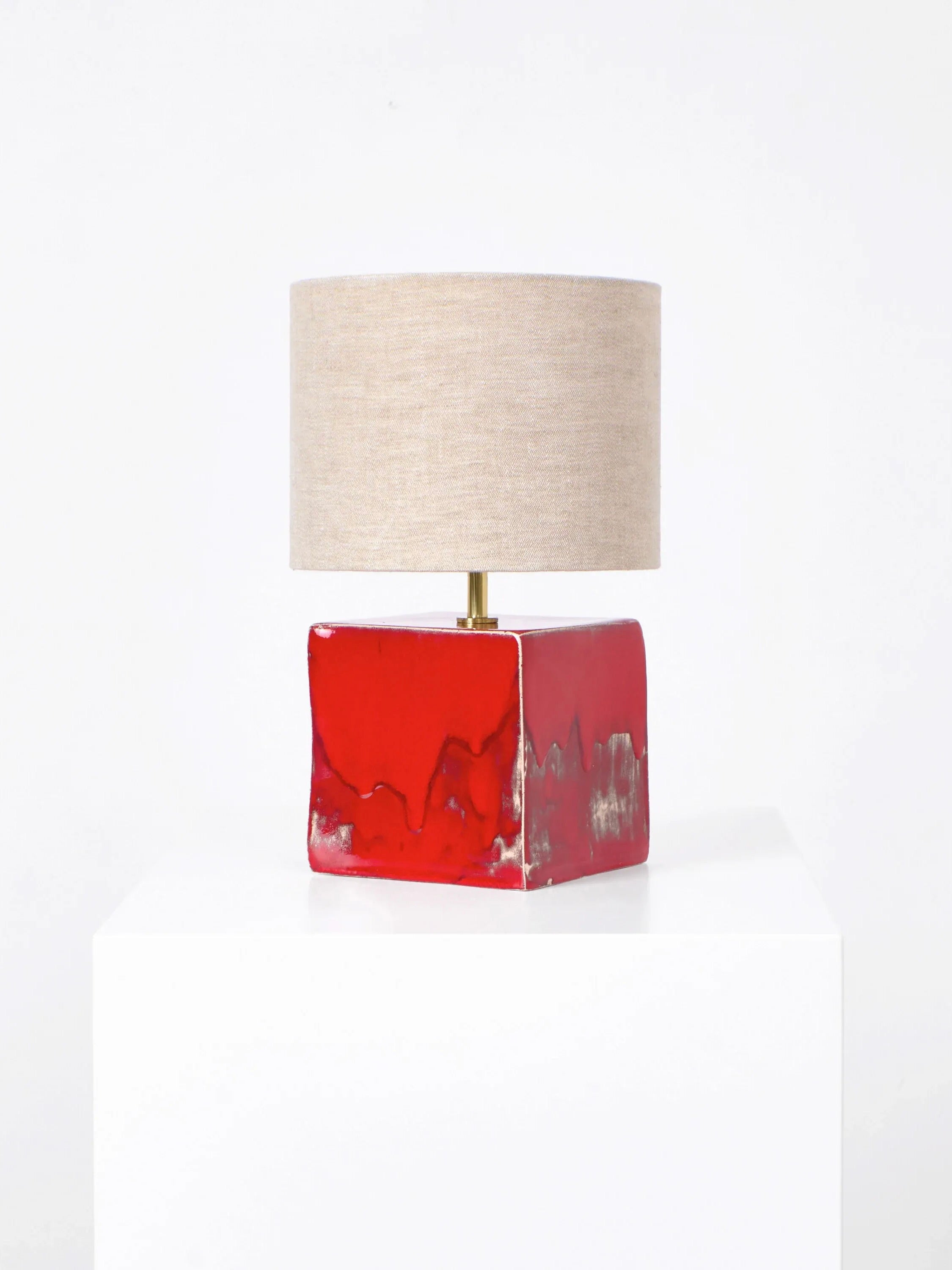 The Arouca Table Light by Project 213A boasts a beige cylindrical shade and a sleek, red glossy rectangular base. Elegantly displayed on a white pedestal against a simple white backdrop, this customizable piece enhances any room's aesthetic.