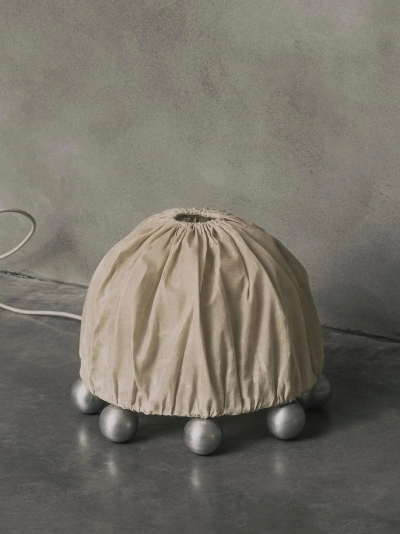 The Kaytar "ALTER" Table Lamp features a beige linen dome top, cinched above six metallic spheres. Set against a textured gray wall, the cord extends across a smooth gray surface, fusing balance and design in this sculptural piece made of aluminium and beeswax.
