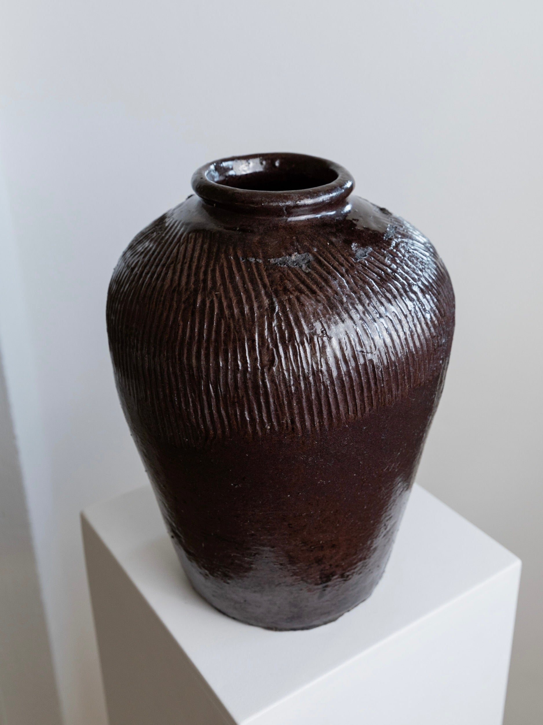 A Bottega Jacobs Antique Chinese 19th Century Ceramic vase with its rustic brown finish and vertical ridges stands elegantly on a white pedestal against a light gray backdrop. This decorative piece features a round shape and a narrow opening at the top, evoking the timeless charm of an antique Chinese rice wine pot.