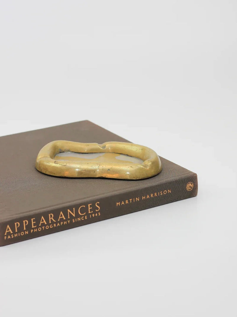 The book "Appearances: Fashion Photography Since 1945" by Martin Harrison rests on a white surface, accompanied by the Brutalist Ash Tray from Boga Avante Shop, featuring a rustic brass finish in an abstract shape.
