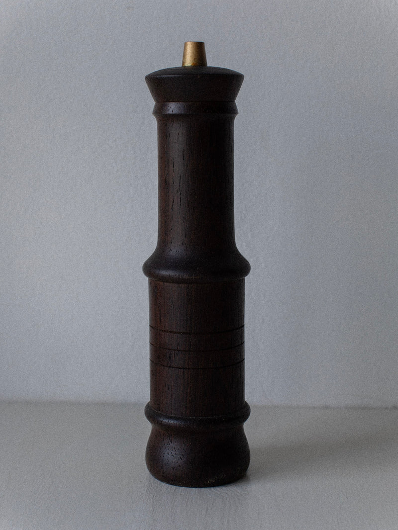 The Wenge Wood Grinder by Out For Lunch stands tall against a light gray background, showcasing its dark brown wenge wood body and golden top knob. The pepper mill, with smooth, slender, and slightly rounded contours, features engraved horizontal lines in the middle section. A vintage grinder of Danish design.
