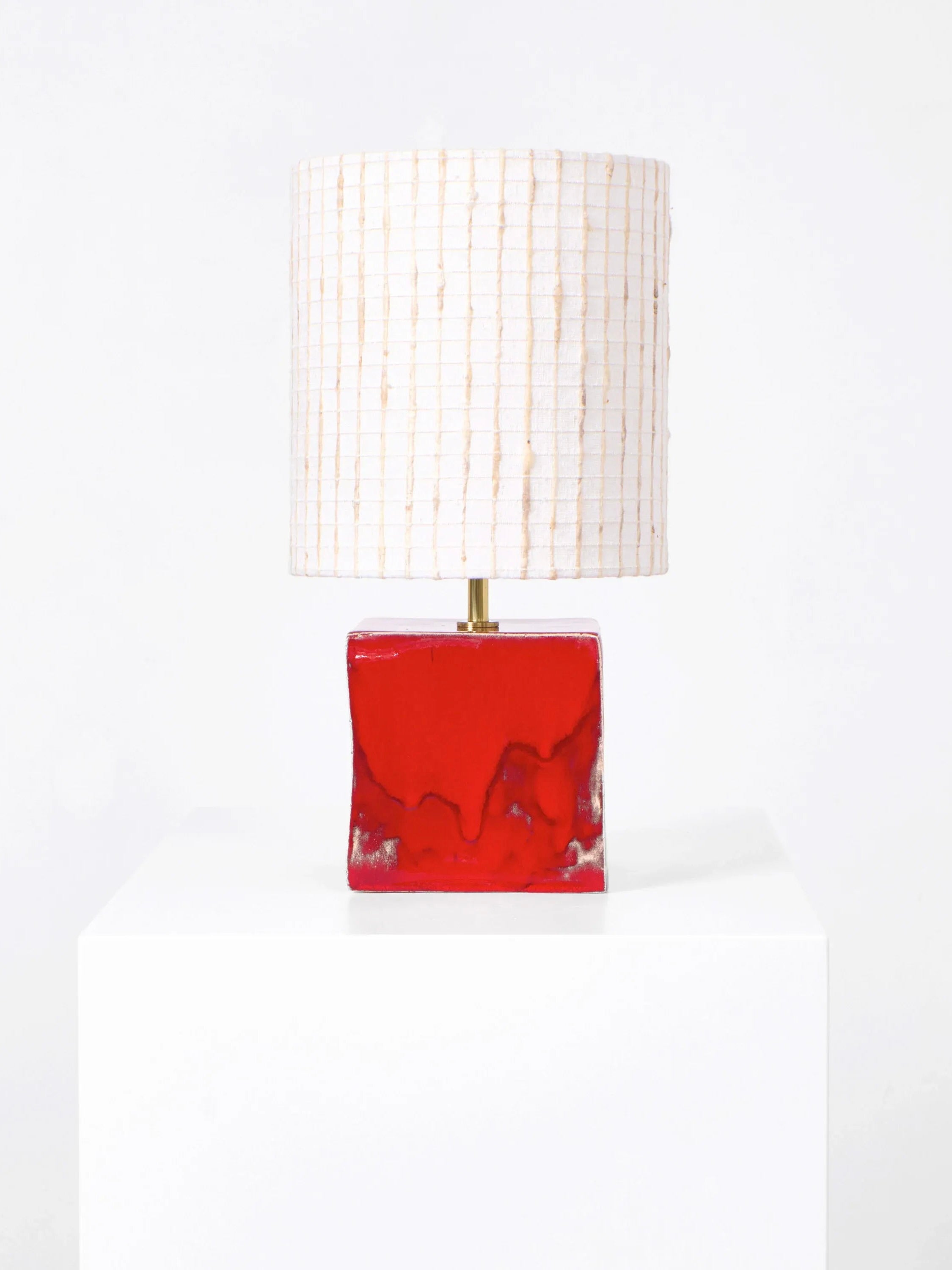 The Arouca Table Light by Project 213A showcases a red, glossy, rectangular ceramic base with a white and beige grid-patterned cylindrical shade, displayed on a white pedestal against a plain white background.