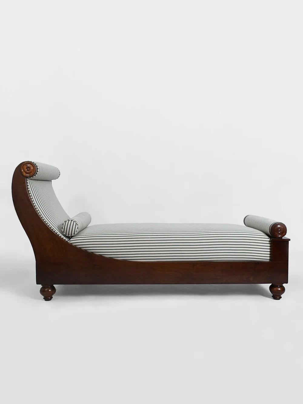 Side view of the Neoclassical Daybed, 19th Century by Barracuda Interiors, showcasing its elegant dark wood frame and white-and-gray striped upholstery. The design includes rolled armrests and a curved back, evoking 19th-century French aesthetics, set against a plain white background.