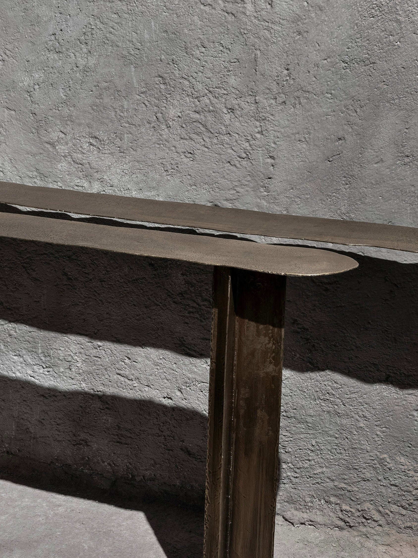 The Muisca Console Table by Ombia Studio features a minimalist design with a flat, elongated surface supported by vertical elements, casting a shadow on a textured gray wall and complemented by brass touches for a simple, modern aesthetic reminiscent of Muisca artifacts.