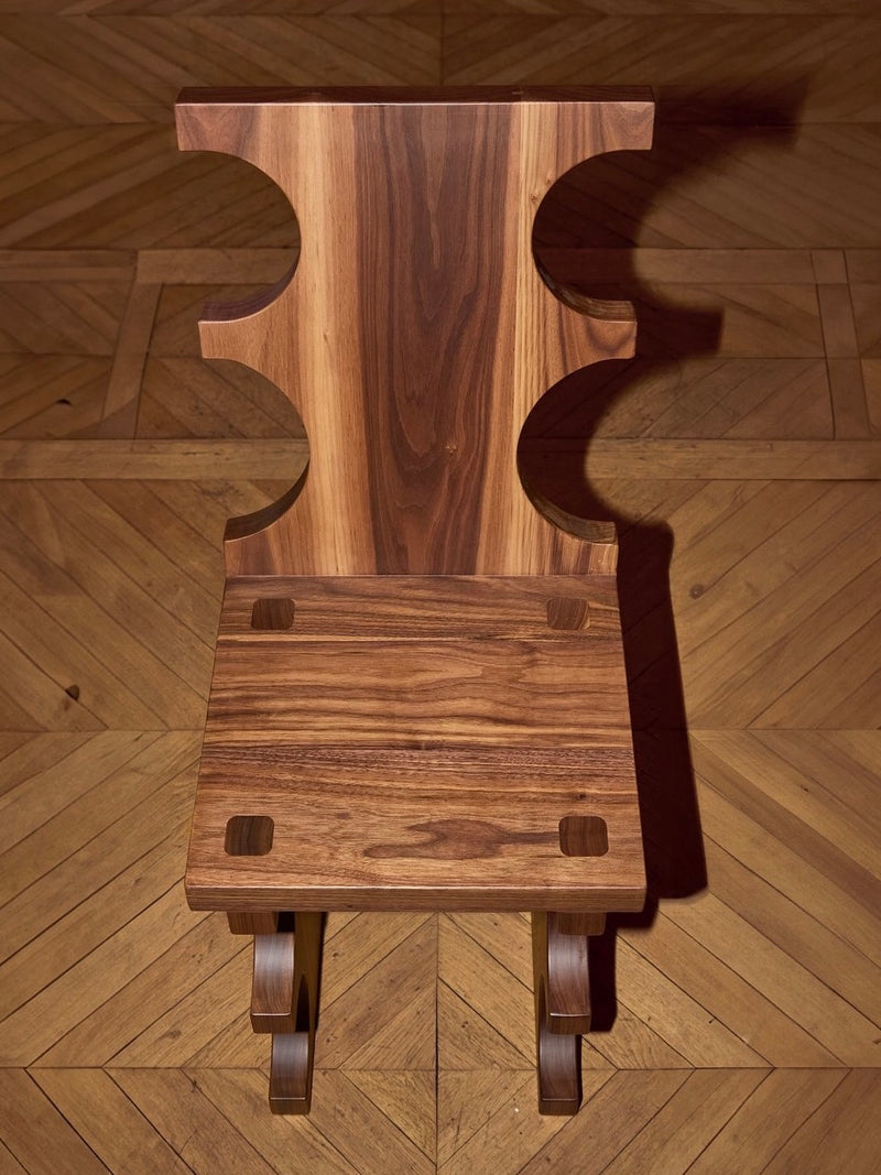 The Portugal Chair No 7, by Project 213A, is a masterfully crafted piece created by skilled artisans from solid walnut. This distinctive chair showcases a unique design with cut-out shapes on the backrest and four square holes on the seat. It features an exquisite warm wood grain finish and looks stunning when placed on a herringbone patterned wooden floor.
