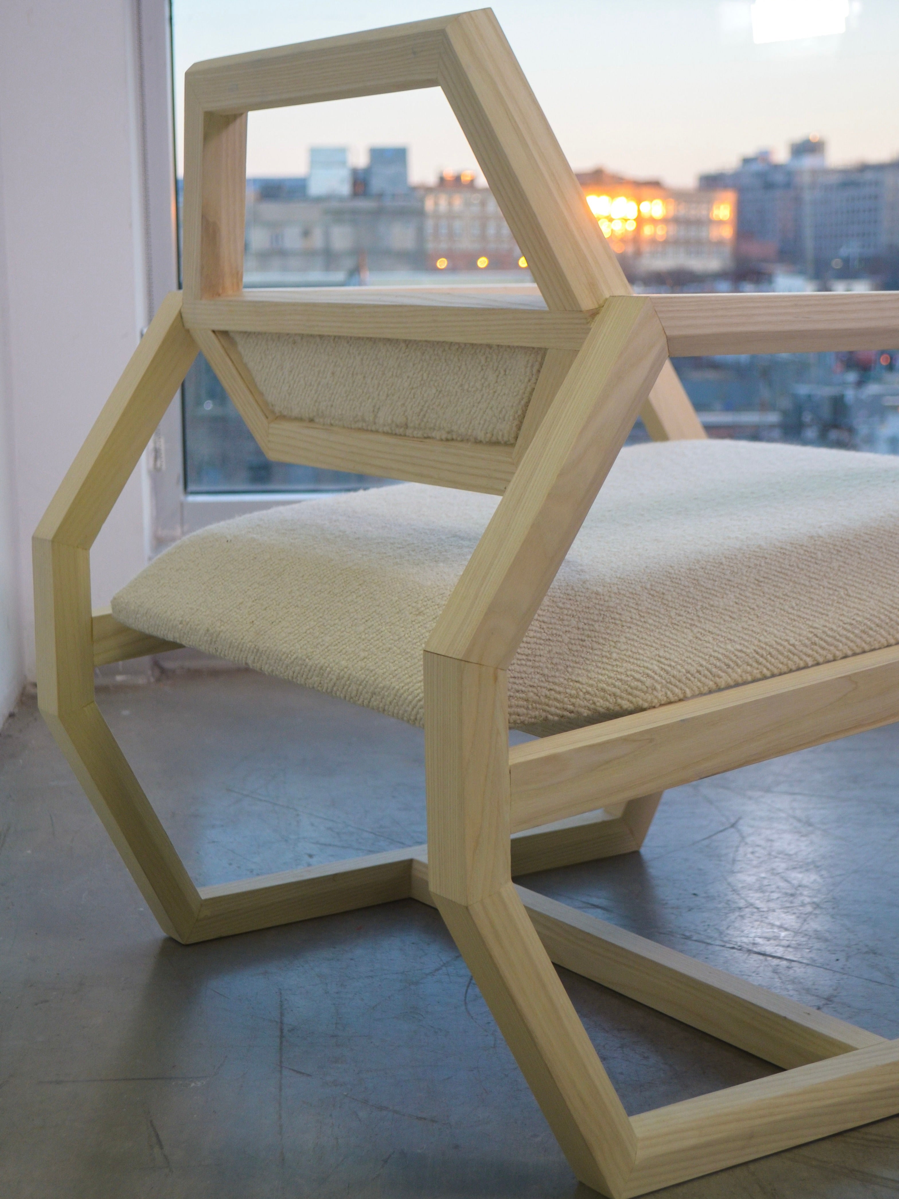 A Marcel Accent Chair from Nathan Chintala Design LLC, showcasing a contemporary wooden frame with a geometric hexagonal design, slanted backrest, and beige woven seat, is situated in a minimalist room with a large window framing an urban skyline at dusk.