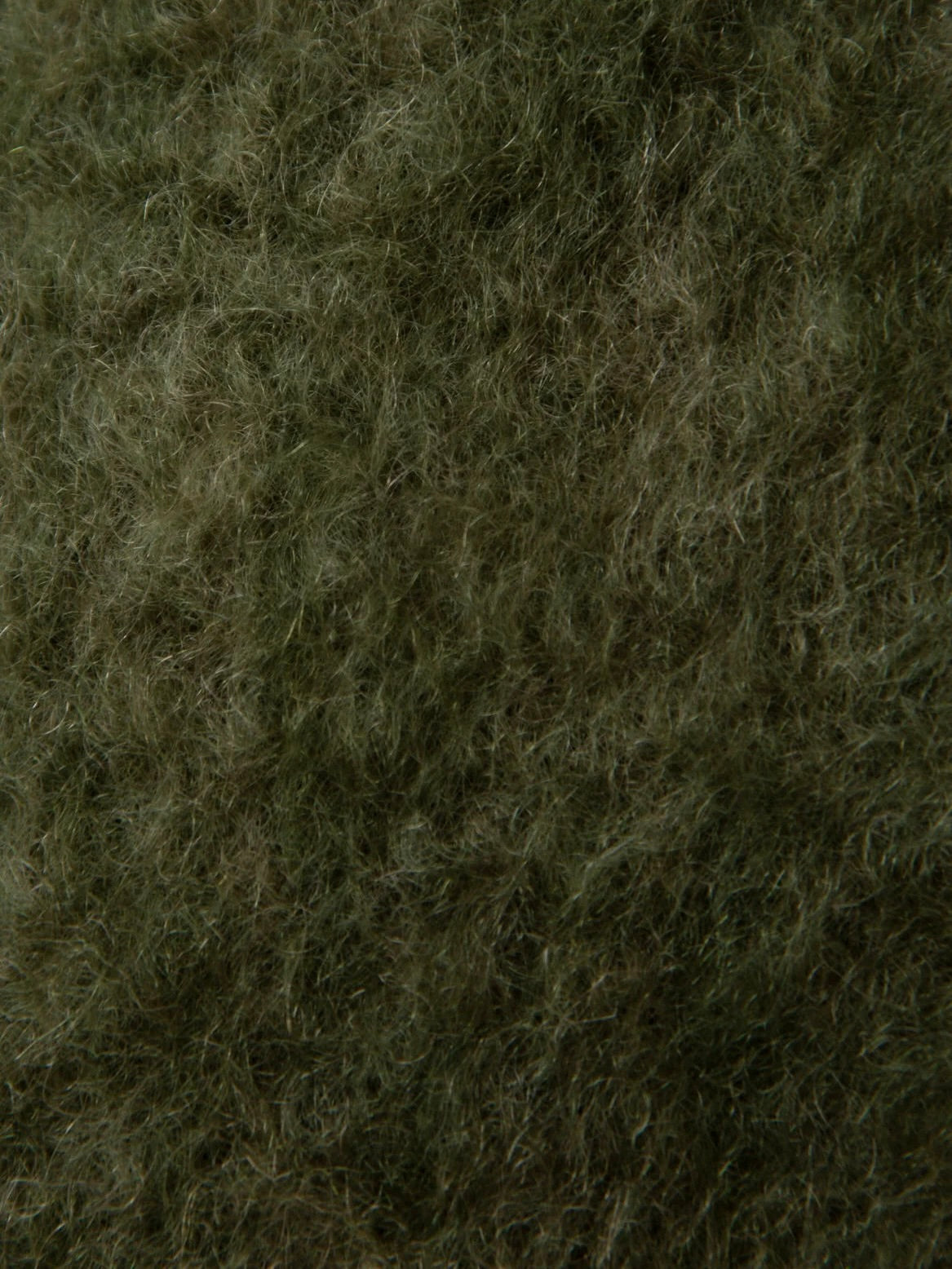 The close-up texture of The Wren's "Arbor no. 2" Throw, with its green, fuzzy, wool-like fabric, resembles a luxurious mohair blanket due to its soft and slightly tangled appearance. The dense interwoven fibers provide the throw with a naturally warm and eco-friendly look.